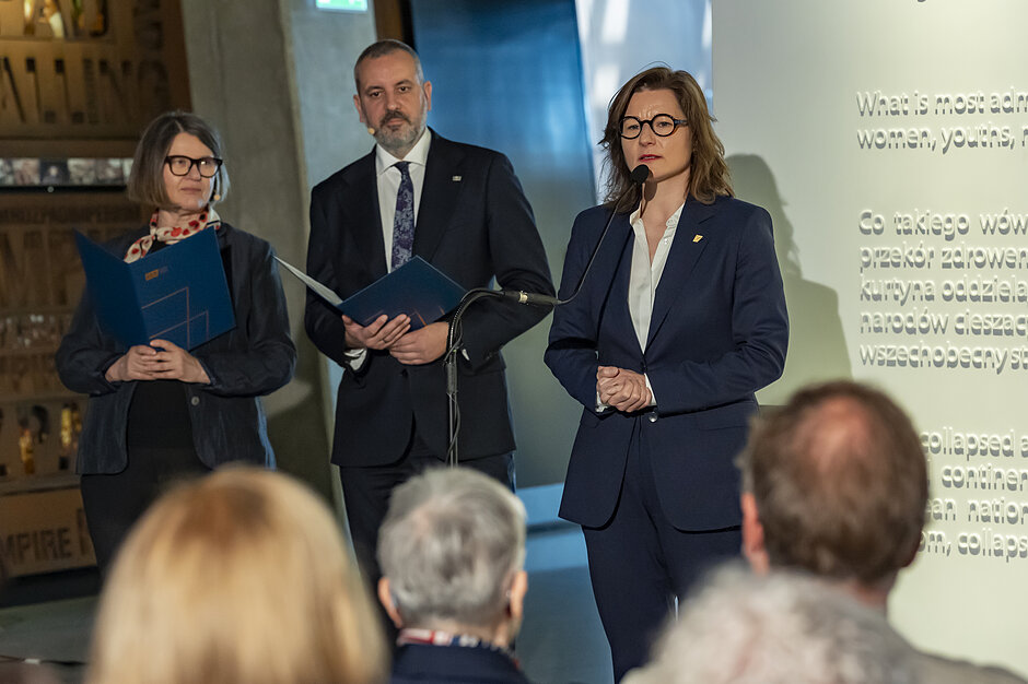Twoje słowa: opisz w 2 zdaniachZdjęcie przedstawia oficjalne spotkanie lub konferencję, podczas której kobieta w ciemnoniebieskim garniturze i okularach przemawia do zgromadzonej publiczności przez mikrofon. W tle po lewej stronie stoją dwie inne elegancko ubrane osoby trzymające foldery, a po prawej widoczny jest fragment wyświetlanej prezentacji z tekstem w języku polskim i angielskim.