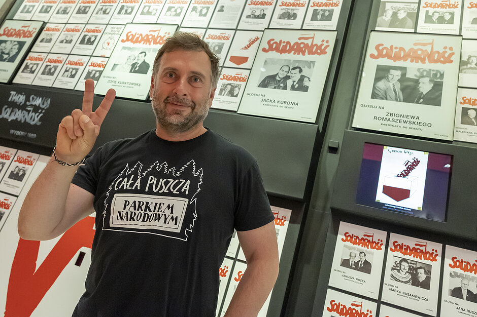 mężczyzna w średnim wieku, prawą dłonią wykonuje gest zwycięstwa, literę V, za nim w tle historyczne plakaty wyborcze solidarności z 1989 roku