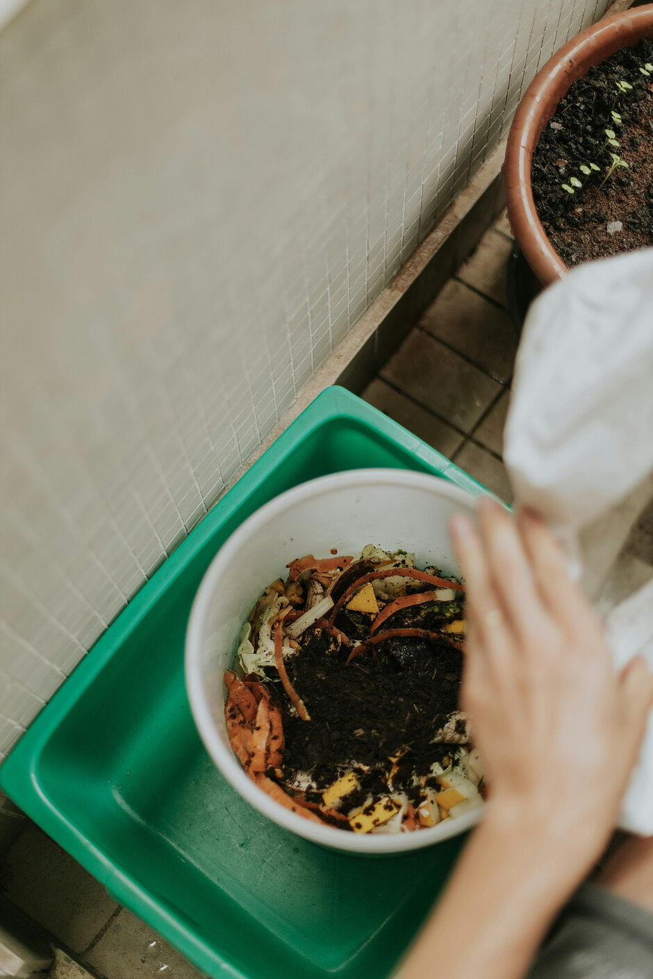 Zdjęcie przedstawia osobę, która wsypuje organiczne resztki oraz ziemię do białej miski umieszczonej w zielonym pojemniku na balkonie. Proces ten wygląda na przygotowywanie domowego kompostu lub pielęgnację roślin w warunkach miejskich.