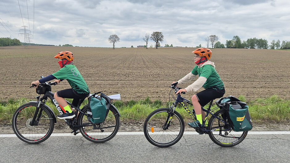 Zdjęcie przedstawia dwóch młodych rowerzystów w pomarańczowych kaskach i zielonych koszulkach, którzy jadą poboczem drogi asfaltowej. Chłopcy poruszają się na rowerach górskich wyposażonych w zielone sakwy, a w tle rozciąga się rozległe, zaorane pole pod zachmurzonym niebem.