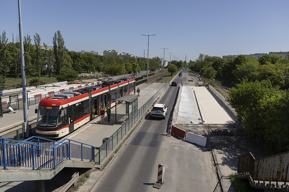 Zdjęcie przedstawia nowoczesny, czerwono-biały tramwaj stojący na przystanku w Gdańsku obok kładki dla pieszych. Po prawej stronie drogi widoczny jest ogrodzony plac budowy, na którym powstaje nowa betonowa konstrukcja infrastruktury miejskiej.