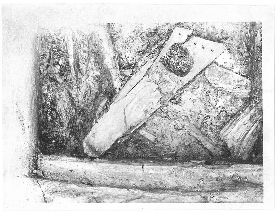 Archiwalne, czarno-białe zdjęcie dokumentuje moment odkrycia tzw. gęśli gdańskich podczas powojennych badań archeologicznych prowadzonych w 1949 roku. Unikalny wczesnośredniowieczny instrument strunowy spoczywa w ziemi pośród reliktów dawnego domostwa, ukazując stan zachowania drewnianego korpusu w chwili odnalezienia
