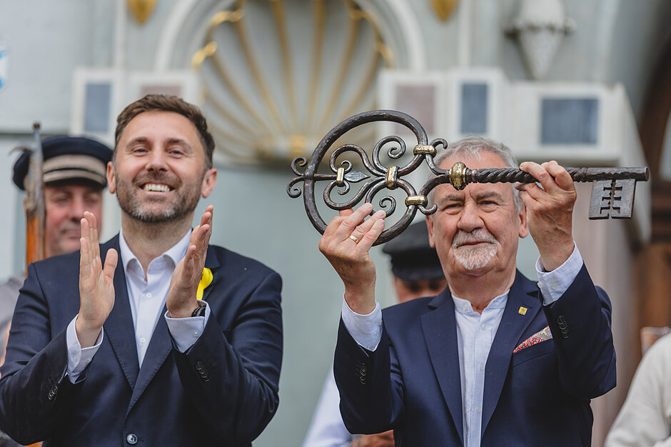 Dwaj mężczyźni, przed Dworem Artusa. Starszy jest po prawej, trzyma na wysokości twarzy duży klucz do miasta. Młodszy jest po lewej, bije brawo 