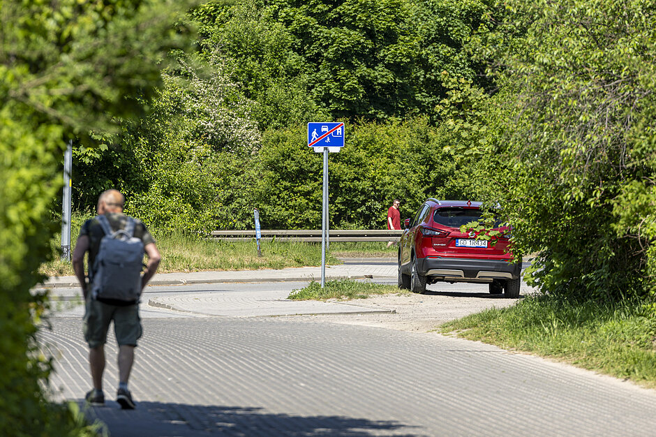 Zdjęcie przedstawia pieszego z plecakiem idącego brukowaną drogą w kierunku czerwonego samochodu marki Nissan. W tle widoczny jest niebieski znak drogowy oznaczający koniec strefy zamieszkania, otoczony gęstą, zieloną roślinnością.
