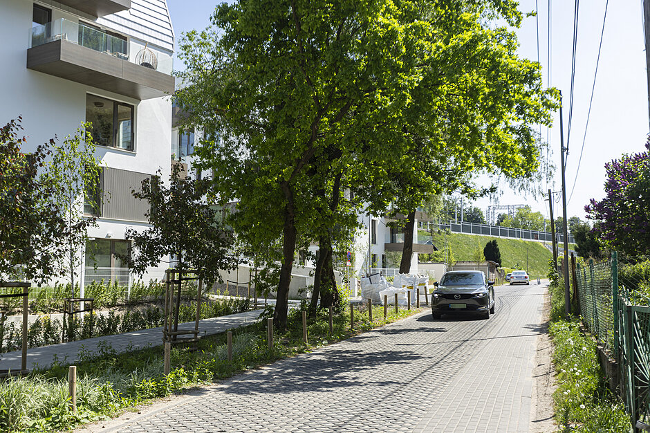 Zdjęcie przedstawia ciemny samochód osobowy jadący wąską, brukowaną uliczką wzdłuż nowoczesnego budynku mieszkalnego. Po bokach drogi widoczne są wysokie, zielone drzewa, trawniki oraz ogrodzenie po prawej stronie.