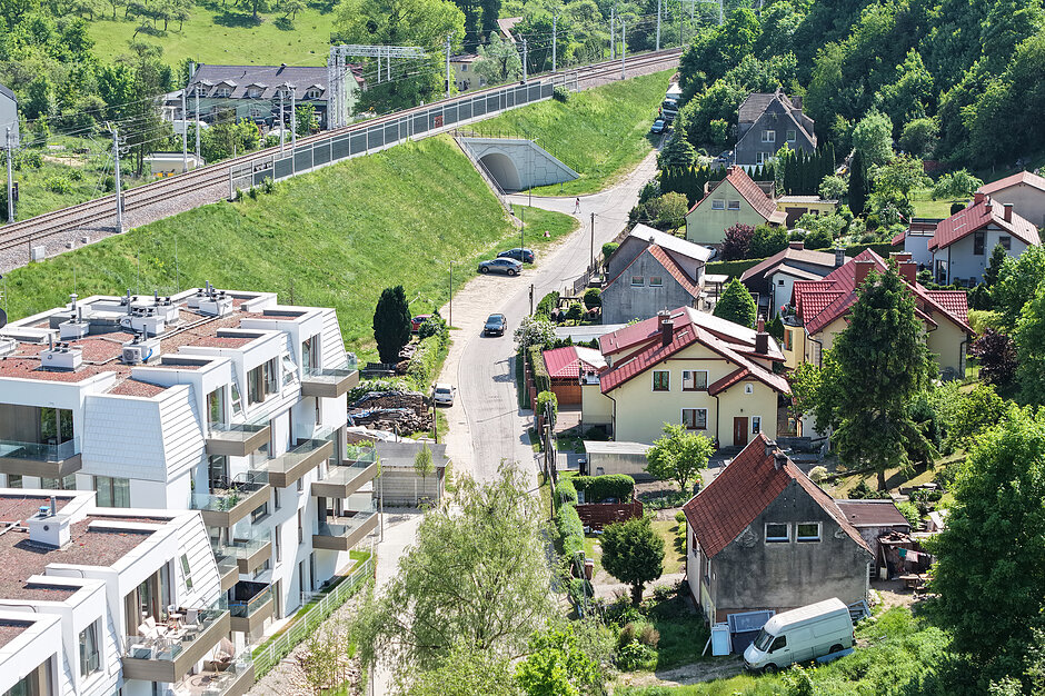 Zdjęcie przedstawia widok z góry na osiedle mieszkaniowe, gdzie nowoczesny, wielorodzinny budynek sąsiaduje z tradycyjnymi domami jednorodzinnymi. W tle widoczna jest linia kolejowa biegnąca na nasypie oraz tunel drogowy otoczony zielonymi wzgórzami.