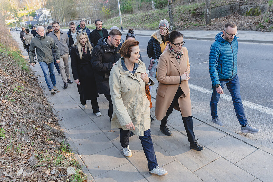Zdjęcie przedstawia grupę ludzi idących chodnikiem wzdłuż asfaltowej drogi. Idący w umiarkowanie ciepłych kurtkach i płaszczach przechodnie poruszają się gęsiego, a w tle widać zbocze z rzadką roślinnością.