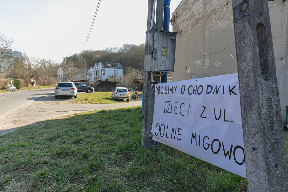 Zdjęcie przedstawia biały baner z odręcznym napisem „PROSIMY O CHODNIK DZIECI Z UL. DOLNE MIGOWO”, przymocowany do betonowego słupa energetycznego. W tle widoczna jest trawiasta przestrzeń, zaparkowane samochody oraz ulica prowadząca w stronę zabudowań i zalesionego wzgórza.