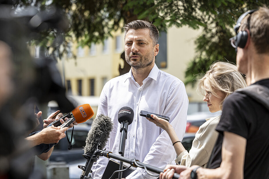 Młody mężczyzna w białej koszuli podczas briefingu. Otaczają go dziennikarze z mikrofonami i kamerami
