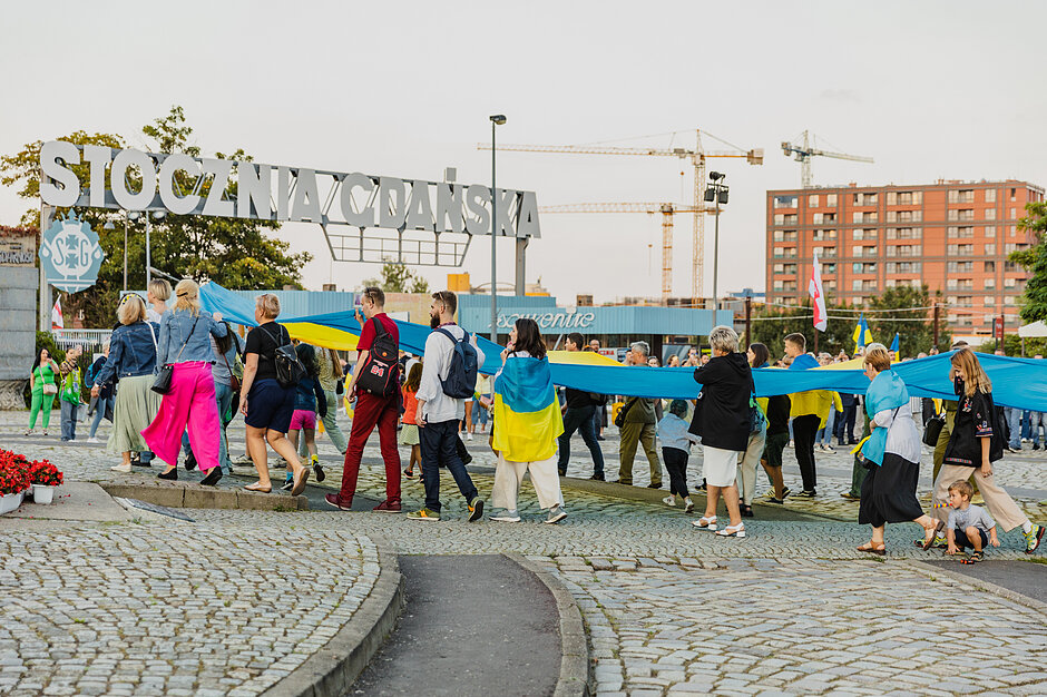 Obchody Dnia Niepodległości Ukrainy w Gdańsku - grupa osób niesie ukraińską flagę. W tle widać Stocznię. 