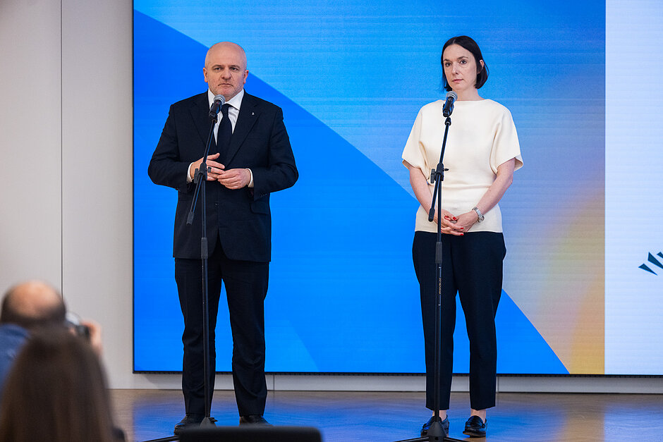 Wiceminister Eliza Zeidler i przewodniczący Rady ds. Współpracy z Ukrainą Paweł Kowal stoją na niebieskim tle podczas konferencji prasowej. Przed sobą mają mikrofony. 