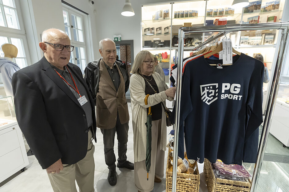 Trójka starszych osób ogląda ubrania i pamiątki w jasnym sklepie z asortymentem sportowym lub uczelnianym. Kobieta przegląda odzież na wieszaku, podczas gdy dwaj towarzyszący jej mężczyźni przyglądają się ekspozycji.