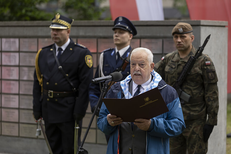 starszy mężczyzna mówi do mikrofonu, za nim stoi żołnierz, strażnik miejski i policjant