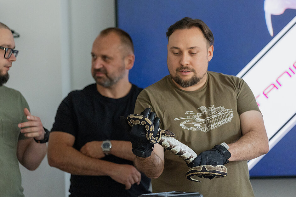 Mężczyzna trzyma węża, mając na rękach rękawice. Obok rozmawia dwóch innych mężczyzn.