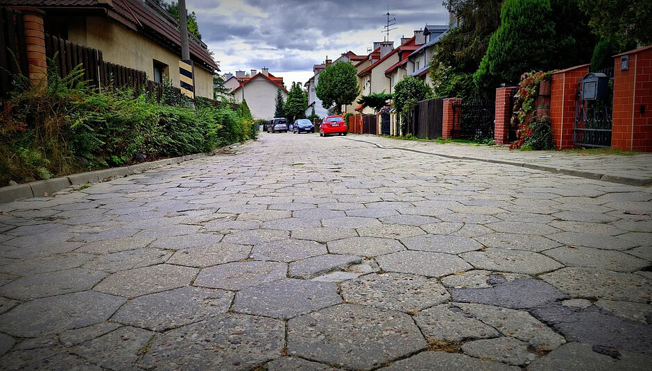 na zdjęciu zbliżenie na nawierzchnię ulicy, w tle kilka budynków mieszkalnych