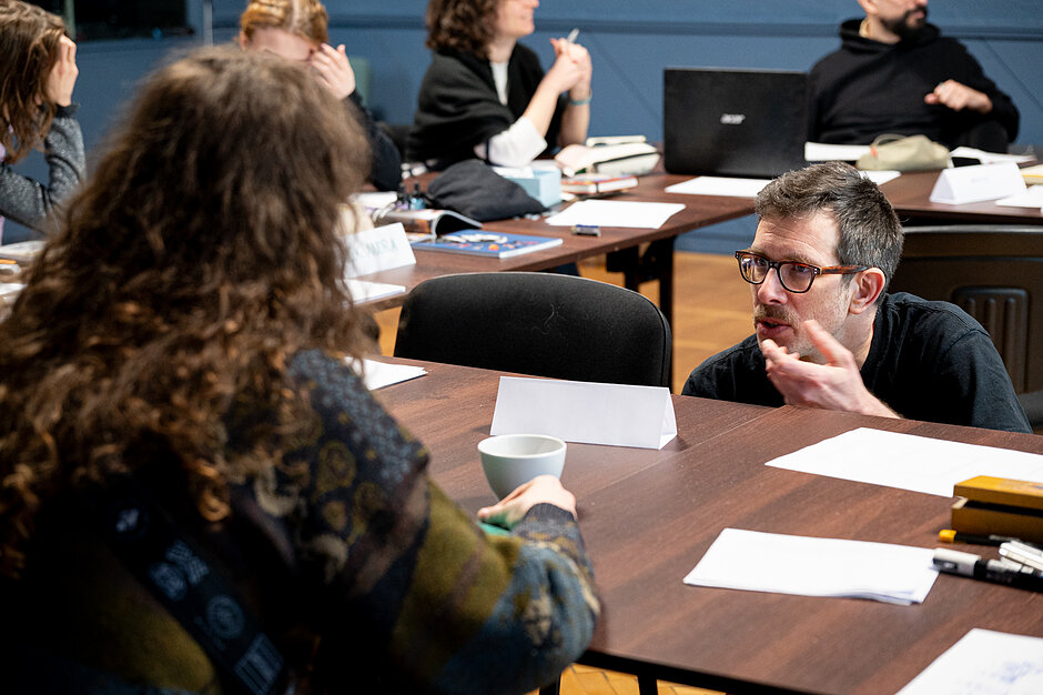 Mężczyzna w okularach żywiołowo rozmawia z kobietą o kręconych włosach przy ciemnym stole konferencyjnym. W tle widać innych uczestników spotkania siedzących przy biurkach z dokumentami i laptopami.