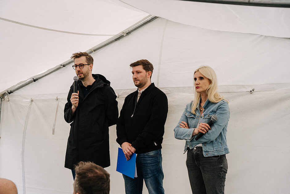 trzy osoby na scenie - dwóch mężczyzn, w tym jeden z mikrofonem oraz kobieta