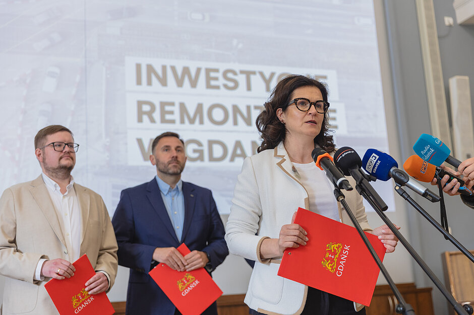 na zdjęciu prezydent gdańska mówi do dziennikarskich mikrofonów, za nią stoją trzej mężczyźni