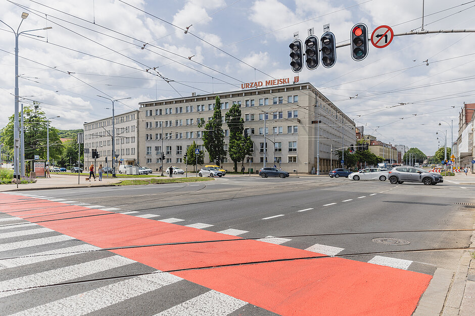 na zdjęciu ulica z wymalowanym przejściem dla pieszych i rowerzystów w tle duży szary budynek z napisem urząd miejski