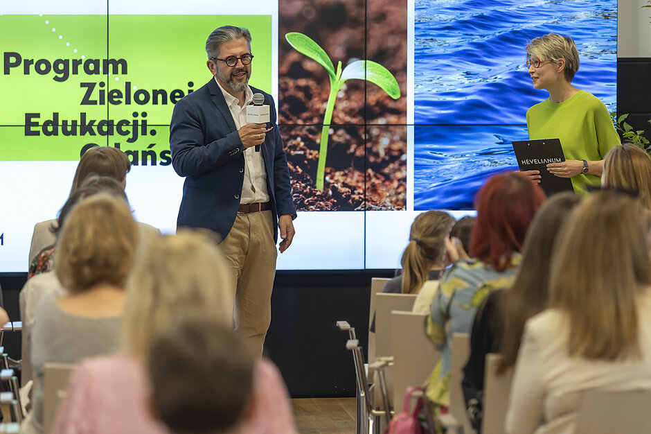 Kobieta i mężczyzna z mikrofonami stoją przed ekranem z prezentacją w sali konferencyjnej.