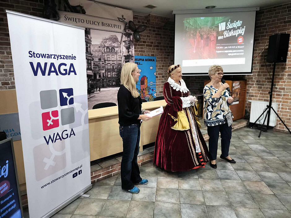 Plansza Stwowarzyszenia WAGA, a obok trzy kobiety