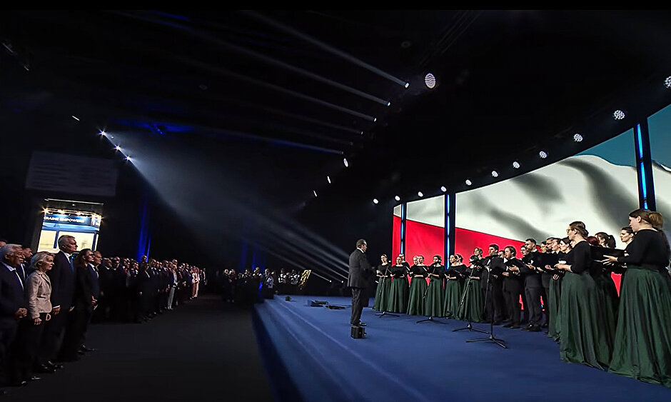 Chór na scenie w wielkiej sali konferencyjnej. Na publiczności stoją ludzie i słuchają hymnu.