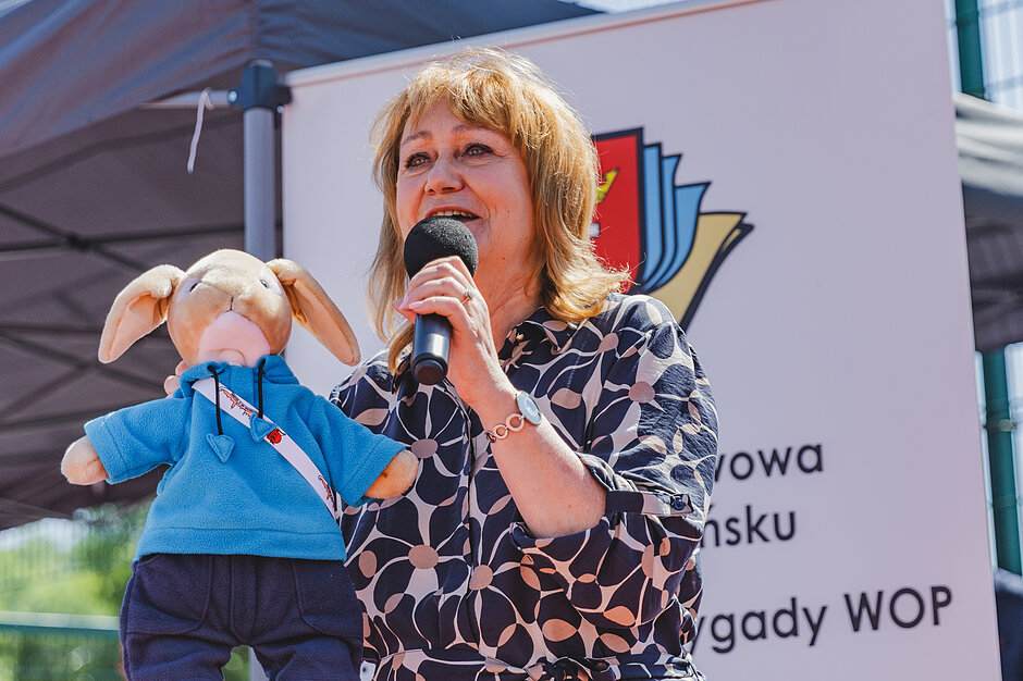 Kobieta trzyma w ręku maskotkę króliczka i mówi do mikrofonu na boisku szkolnym.
