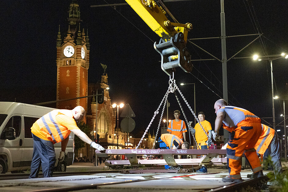 zdjęcie w nocy, widać kilku robotników trzymających dużą stalową konstrukcję unoszoną na dźwigu