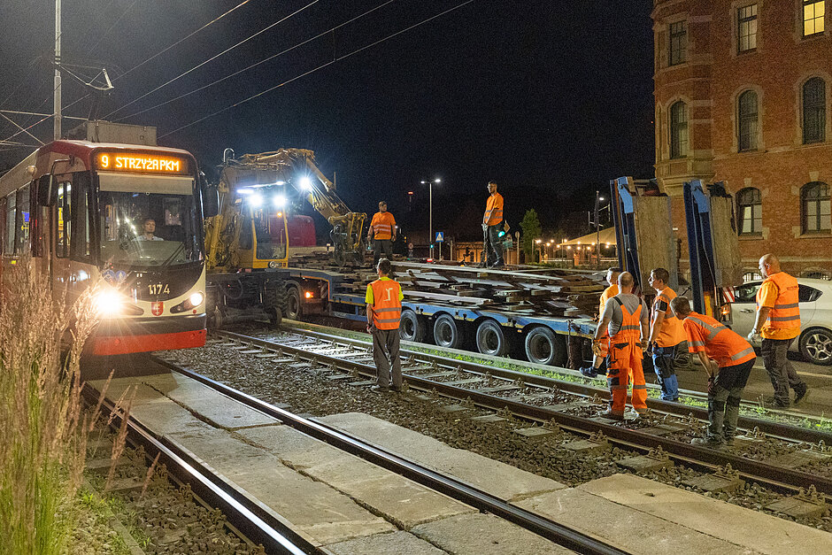 zdjęcie w nocy, widać jadący tramwaj , a po prawej stoją robotnicy w pomarańczowych kombinezonach