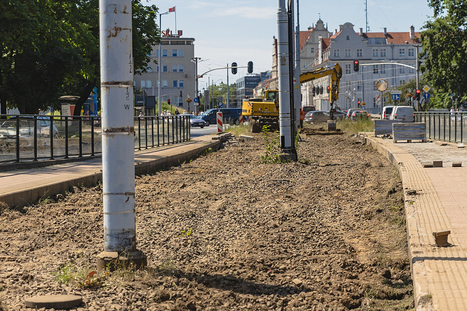rozkopany teren gruntowy, widać słupy sieci trakcyjnej, w tle samochody i budynek urzędu po lewej, 