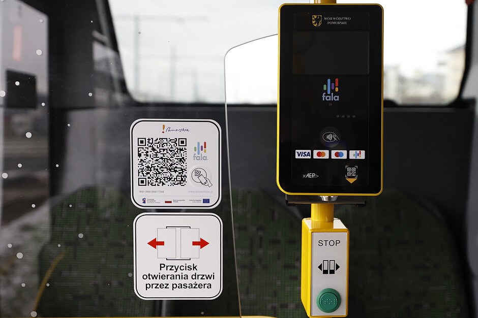 Zdjęcie przedstawia nowoczesny, żółto-czarny czytnik systemu Fala oraz naklejkę z kodem QR, zamontowane w pojeździe komunikacji miejskiej w województwie pomorskim. Urządzenia te służą pasażerom do bezgotówkowego opłacania przejazdów oraz zbliżeniowego kasowania biletów za pomocą karty płatniczej lub smartfona.