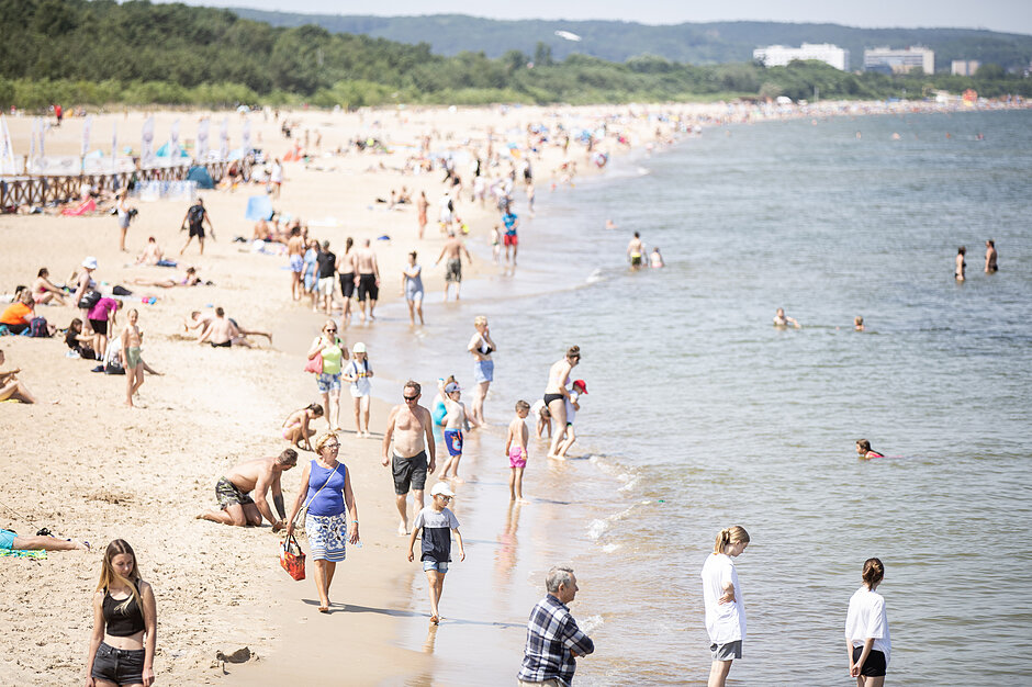 Zdjęcie przedstawia szeroką, piaszczystą plażę nad Morzem Bałtyckim wypełnioną wypoczywającymi ludźmi. Liczni plażowicze spacerują wzdłuż brzegu, opalają się na piasku oraz kąpią się w morzu.