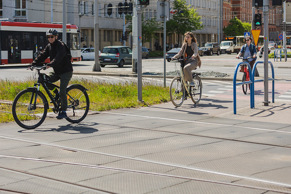 rowerzyści przejeżdżają przez tory tramwajowe, w tle jedzie tramwaj