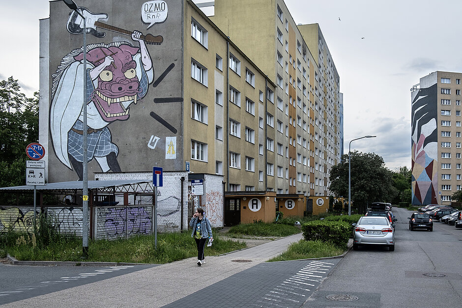 Kobieta idzie chodnikiem przez osiedle i mija mural na bloku. W tle kolejny mural na innym bloku.