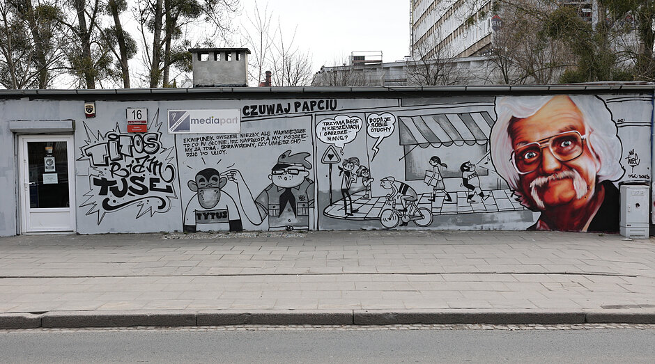 Mural z Papciem Chmielem, Tytusem, Romkiem i Atomkiem na podłużnym niskim budynku w mieście.