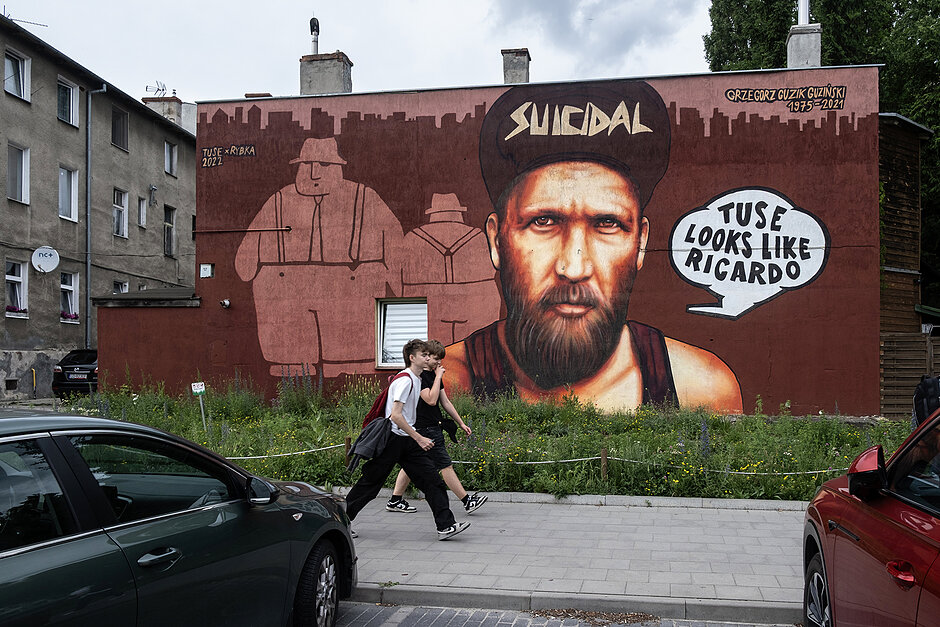 Mural mężczyzny z brodą i w czapeczce na starym budynku w starej dzielnicy miasta. Mijają go dwaj chłopacy.