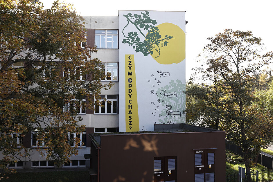 Mural na budynku szkoły z żółtą kulą słońca i przesłaniem antysmogowym.