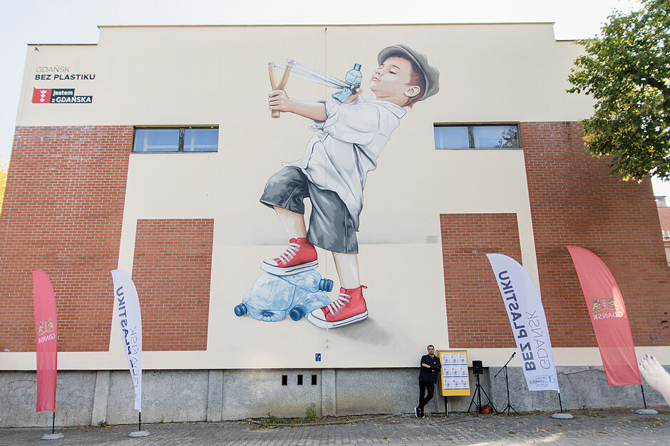 Mural na budynku szkoły z chłopcem strzelającym z procy plastikową butelką.