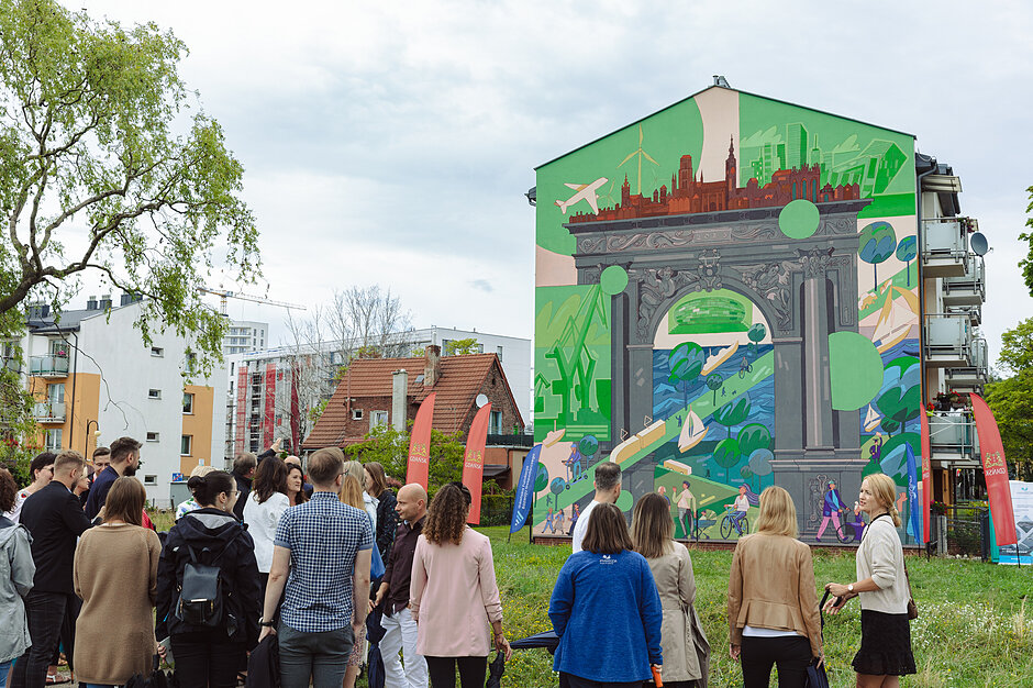 Mural z motywami architektury gdańskiej na budynku mieszkalnym. Podziwia go grupa osób.