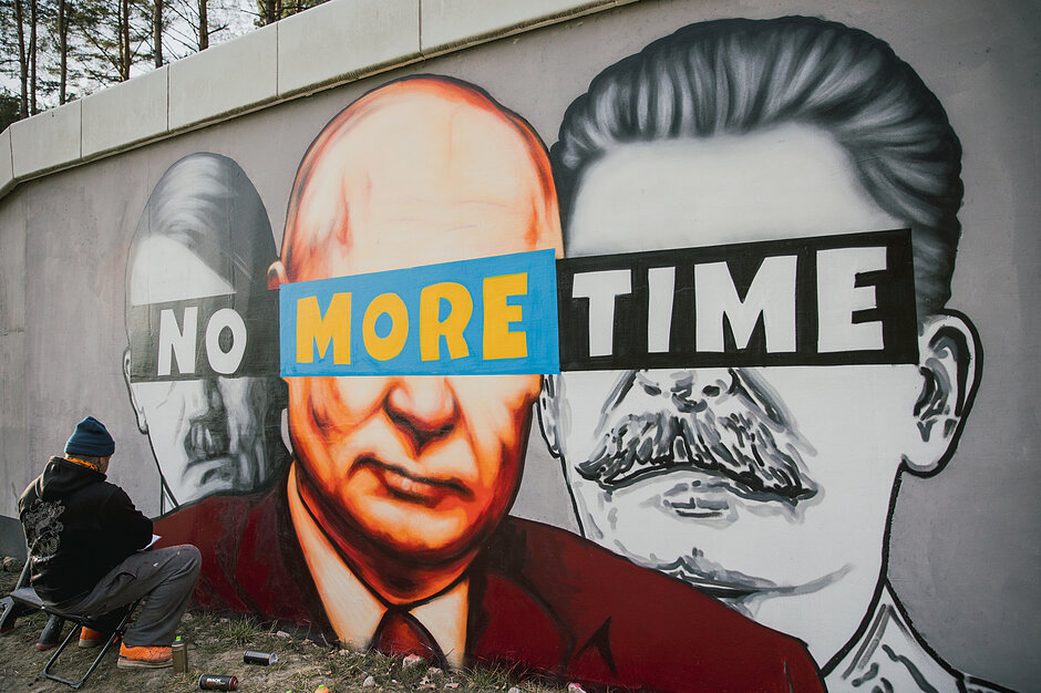 Mężczyzna maluje na murze mural z podobiznami Hitlera, Putina i Stalina, którzy mają zasłonięte oczy napisem No More Time.