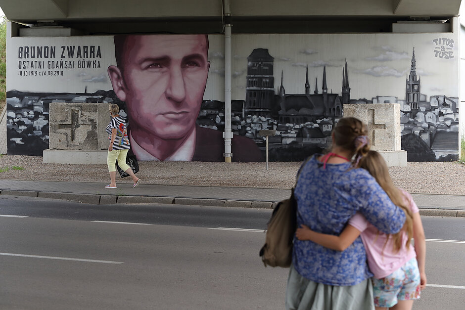 Dwójka obejmujących się ludzi ogląda mural pod wiaduktem z mężczyzną i widokiem Gdańska.