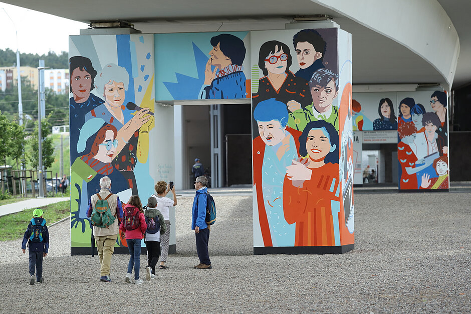 Ludzie oglądają murale na filarach wiaduktu przedstawiające różne kobiety.