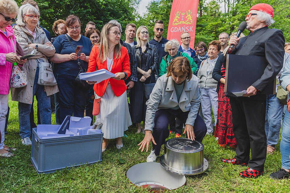 Tłum ludzi zgromadzonych w parku wokół wkopanej w ziemię kapsuły czasu.