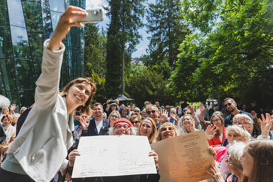 Kobieta trzyma wysoko telefon i robi zdjęcie selfie z tłumem osób za nią w parku.