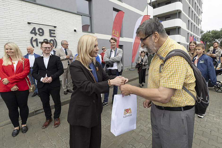 Zdjęcie przedstawia oficjalne spotkanie przed nowym budynkiem z numerem 49 E, podczas którego kobieta w ciemnym garniturze wręcza mężczyźnie białą torbę upominkową z logo Gdańska. W tle zgromadziła się grupa obserwujących wydarzenie osób, a obok widoczne są flagi, w tym czerwona flaga Gdańska.