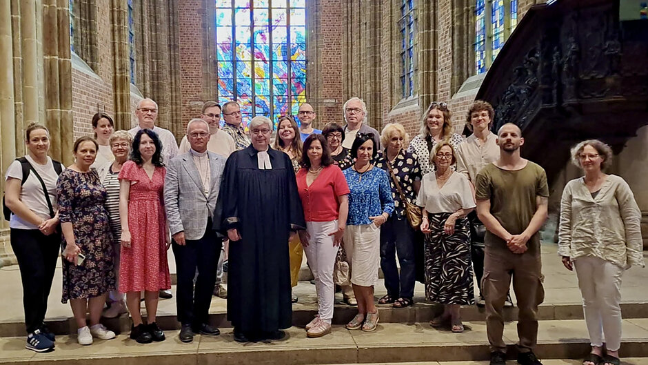 Zdjęcie przedstawia dużą grupę osób pozujących do wspólnej fotografii na schodach we wnętrzu zabytkowego, gotyckiego kościoła. W centrum stoją dwaj duchowni – jeden w tradycyjnej ciemnej todze, a drugi w jasnej marynarce z koloratką – otoczeni przez pozostałych uczestników spotkania ubranych w stroje codzienne i wyjściowe.