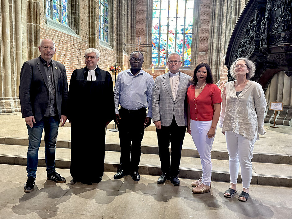 Zdjęcie przedstawia grupę sześciu osób stojących w rzędzie we wnętrzu zabytkowego kościoła o gotyckiej architekturze z widocznym w tle witrażem. Wśród zgromadzonych znajduje się duchowny w tradycyjnej tozdze, a pozostali uczestnicy spotkania ubrani są w stroje wyjściowe oraz casualowe.