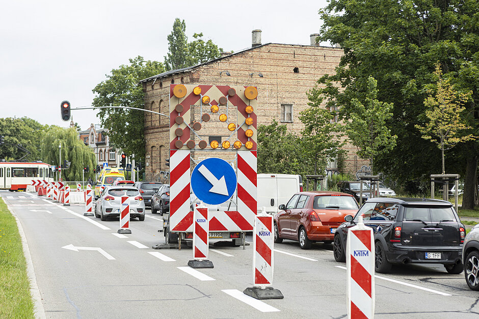 na zdjęciu ulica, widać na prawym pasie sznur stojących samochodów, po środku stoi znak drogowy ze strzałką