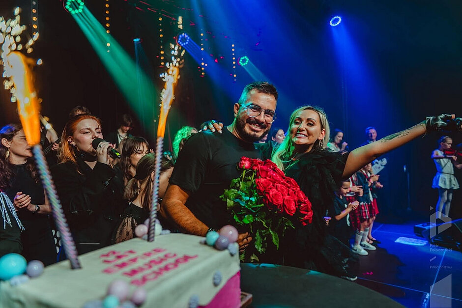 Kolorowe zdjęcie wykonane na scenie podczas wydarzenia artystycznego lub jubileuszowego. Na pierwszym planie widoczny jest duży tort z zapalonymi fontannami iskier. W centrum kadru dwie osoby pozują do zdjęcia, obejmując się ramionami. Jedna z nich trzyma okazały bukiet czerwonych róż. Wokół znajduje się grupa uczestników wydarzenia, w tym osoby z mikrofonami i instrumentami. Scenę oświetlają intensywne niebieskie, zielone i fioletowe reflektory, a w tle widać występujących oraz publiczność lub uczestników zgromadzonych na scenie. Zdjęcie oddaje atmosferę uroczystego świętowania i wspólnego występu.