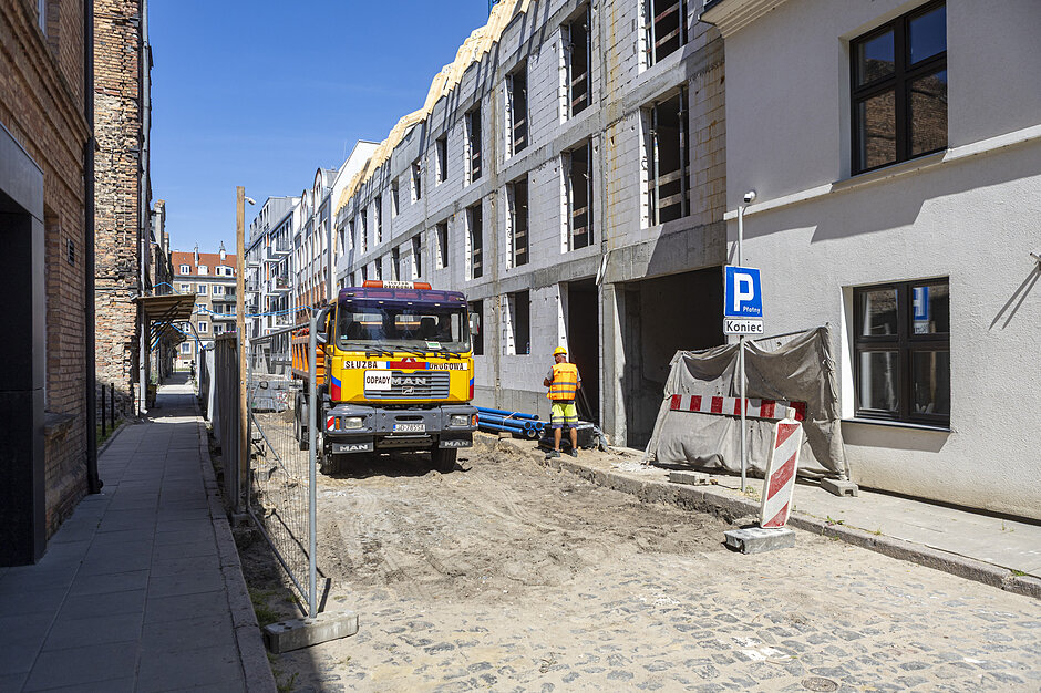 Fotografia przedstawia plac budowy nowego budynku wielorodzinnego zlokalizowanego wzdłuż wąskiej, brukowanej ulicy miejskiej. Na ogrodzonym terenie stoi pomarańczowa ciężarówka marki MAN, obok której pracuje robotnik w odblaskowej kamizelce i kasku ochronnym.